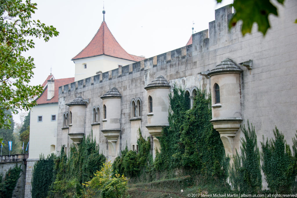 Long crenellated castle wall with repeated projecting window bays, ivy, and a red-roofed tower behind.