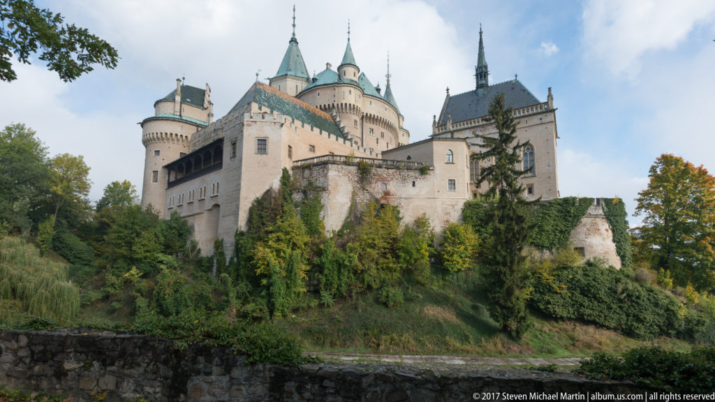 Bojnice Castle towers and steep roofs rise above trees, retaining walls, and the water of the moat.
