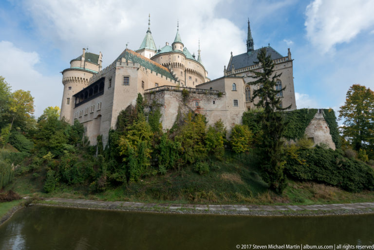 Wide view of Bojnice Castle across the moat, with clustered towers, steep roofs, gallery, and wooded bank.