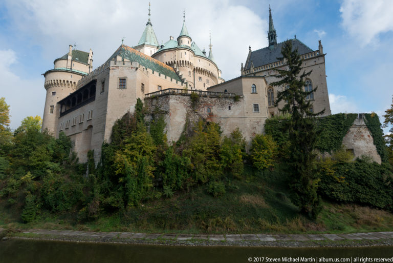 Bojnice Castle above a curved moat, with ivy-covered walls, trees, towers, and steep roofs.