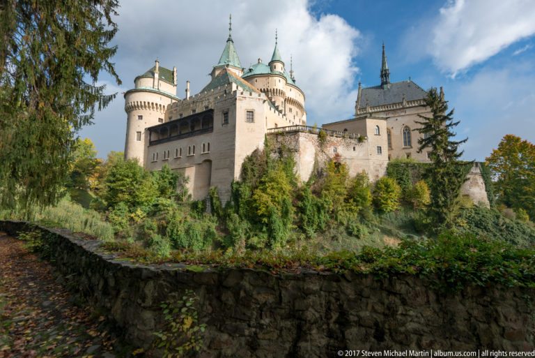 Sunlit Bojnice Castle above a dark stone wall, with clustered towers and autumn foliage.