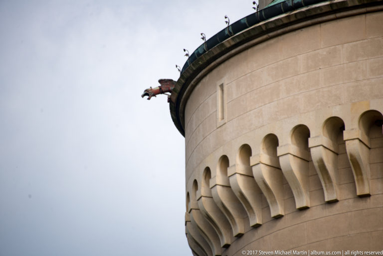 Dragon-shaped metal waterspout projects from a round tower above a band of large stone corbels.
