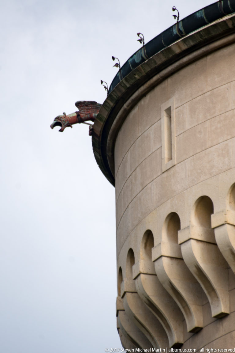 Close upward view of a dragon-shaped metal waterspout on a round tower with stone corbels.