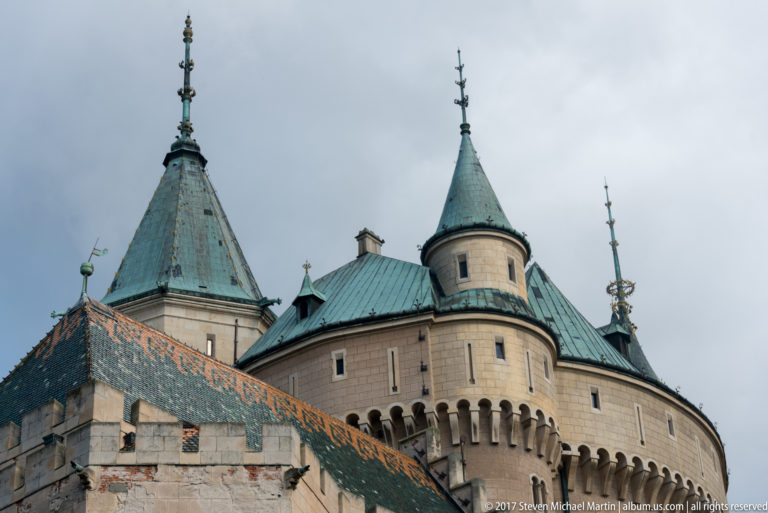 Clustered Bojnice Castle towers with green metal roofs, corbelled walls, finials, and a steep patterned tile roof.