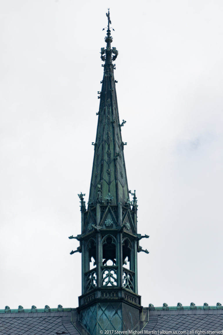 Tall openwork metal spire with Gothic-inspired tracery and small projecting ornaments against the sky.