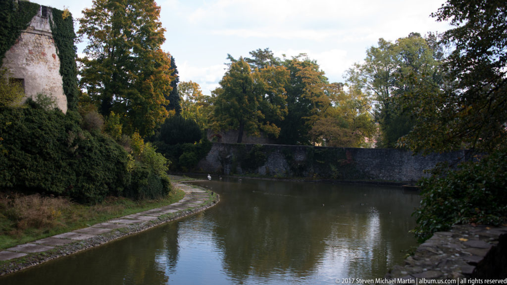 Curving moat bordered by stone edging, mature trees, and a masonry wall in the Bojnice Castle grounds.