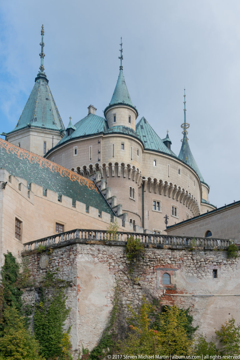 Pale round towers with green roofs and corbelled upper walls rise above a lower masonry perimeter wall.
