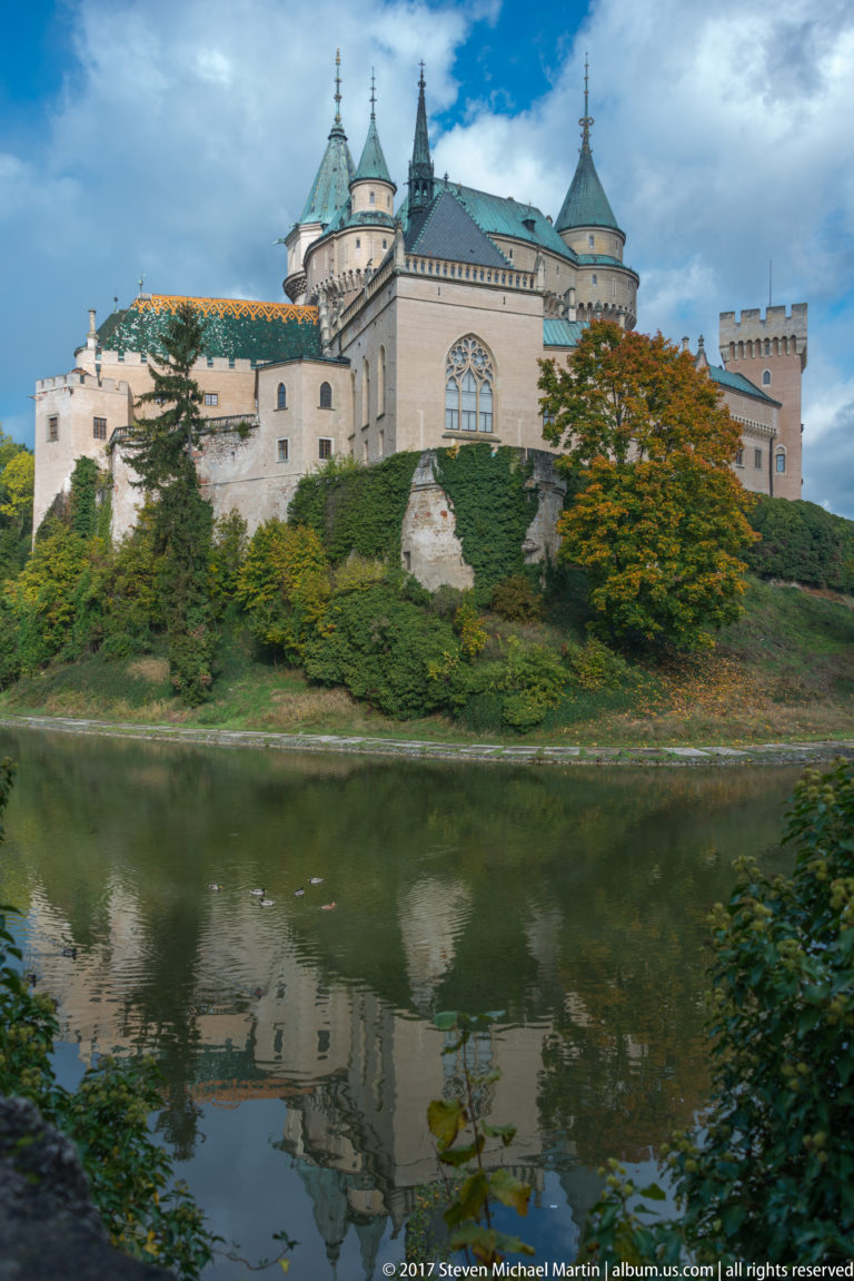 Wide view of Bojnice Castle across the moat with pale towers, steep roofs, autumn trees, and water reflections.
