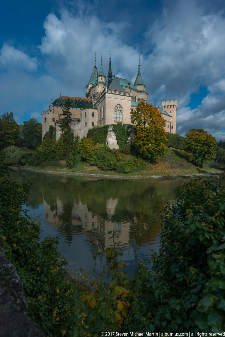Bojnice Castle reflected in the moat beneath dark clouds, with autumn foliage and dense shrubs along the bank.