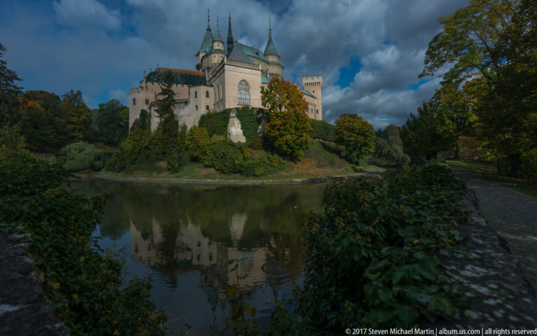 Bojnice Castle seen across the moat from a shaded path with shrubs, reflections, and heavy clouds.