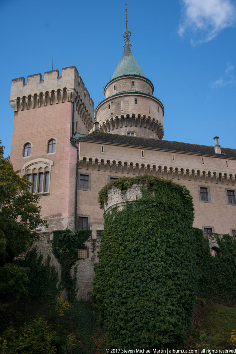 Round corbelled tower with green roof beside a crenellated square tower and long pale wing at Bojnice Castle.