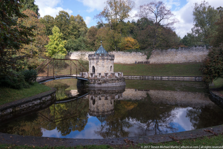 Small polygonal stone turret with metal roof and footbridge reflected in the moat beside a long stone wall.