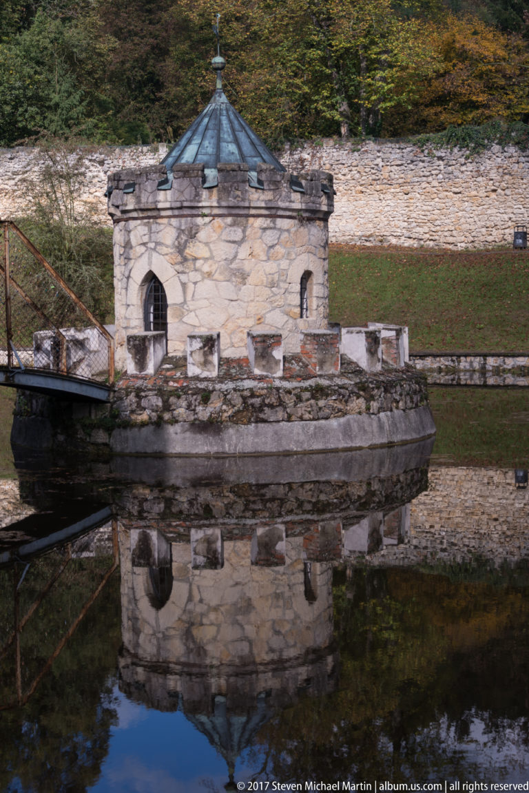 Close view of a small stone moat turret with arched doorway, narrow opening, metal roof, bridge, and reflection.