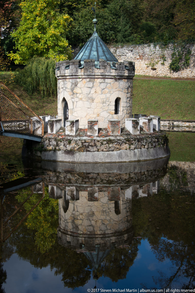 Stone moat turret with small opening and metal roof reflected in calm water before a long boundary wall.