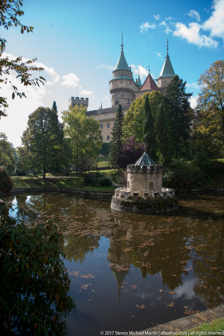 Small moat turret reflected in the water with Bojnice Castle rising behind mature trees.