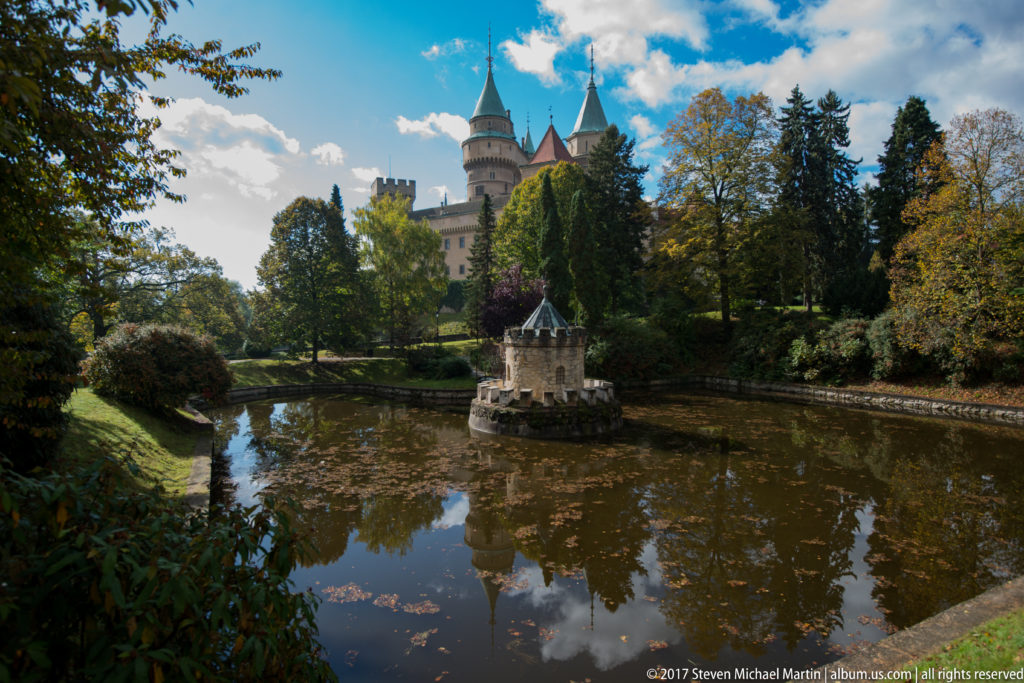 Wide view of the moat turret and Bojnice Castle framed by autumn trees and reflected clouds.