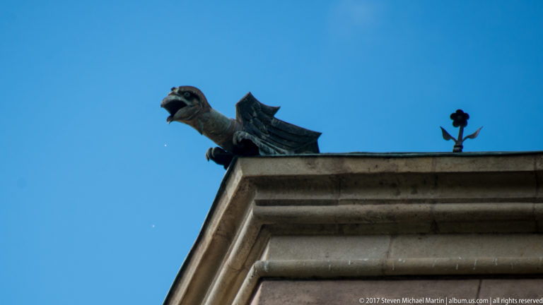 Dragon-shaped metal gargoyle with open mouth and wings projecting from a stone roof corner against blue sky.