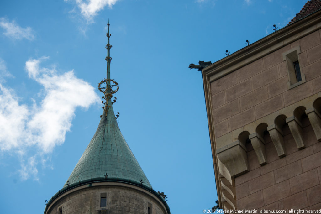 Green conical tower roof with elaborate metal finial beside a pale corbelled tower wall.