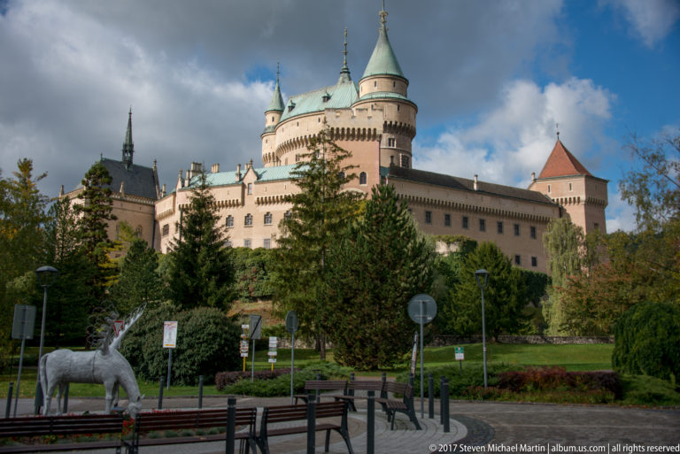 Bojnice Castle above trees and visitor grounds, with clustered towers, signs, benches, and a white animal sculpture in front.