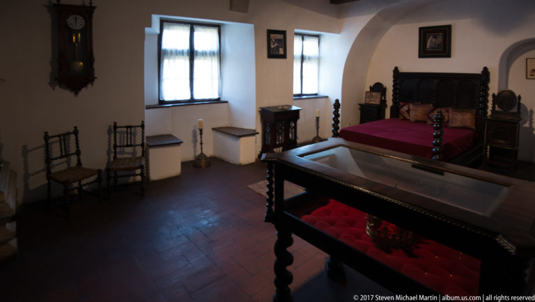 Historic bedchamber with carved dark wood bed, red coverlet, chairs, cabinets, and recessed windows.