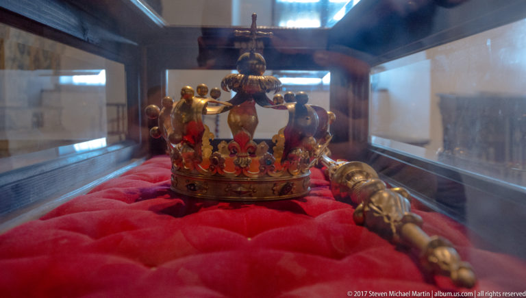 Jeweled crown and ceremonial scepter displayed on red fabric inside a glass case.