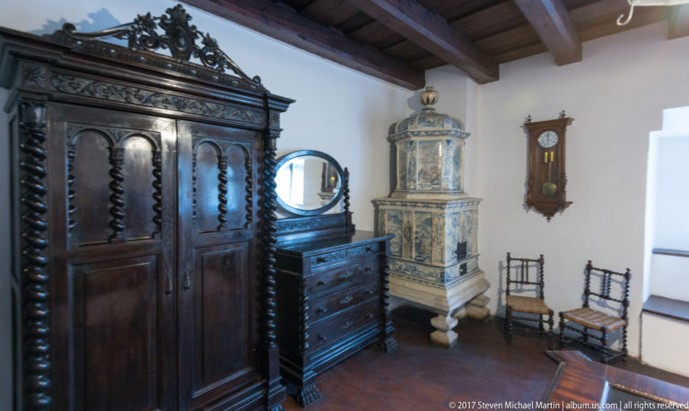 Dark carved wardrobe and dresser beside a pale tiled stove, wall clock, and two chairs.