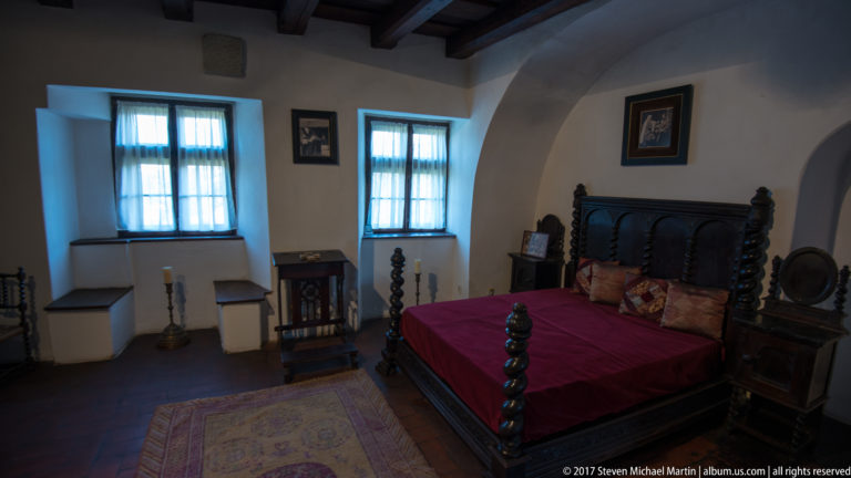 Carved wooden bed with red coverlet centered between recessed windows in a historic bedchamber.