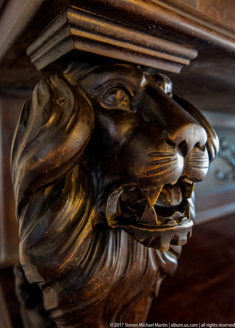 Close view of a carved wooden lion head projecting from the corner of a dark furnishing.