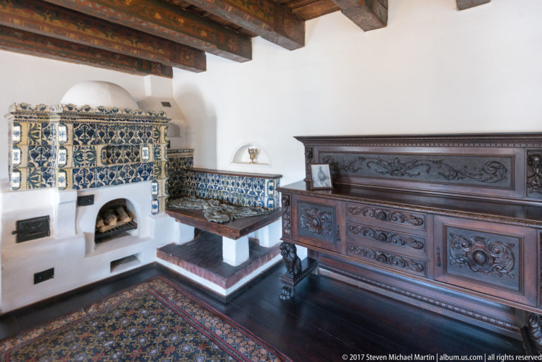 Room corner with blue-and-white tiled stove and bench, carved chest, timber beams, and patterned rug.