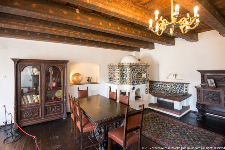 Dining room with long wooden table, chairs, carved cabinets, tiled stove, and painted ceiling beams.