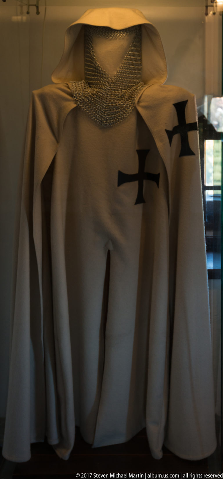 White displayed garments with black cross motifs and a broad woven head covering behind glass.