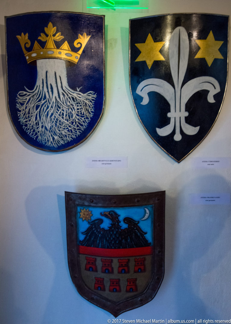 Painted heraldic shields featuring floral, eagle, star, and divided-field motifs on a white wall.