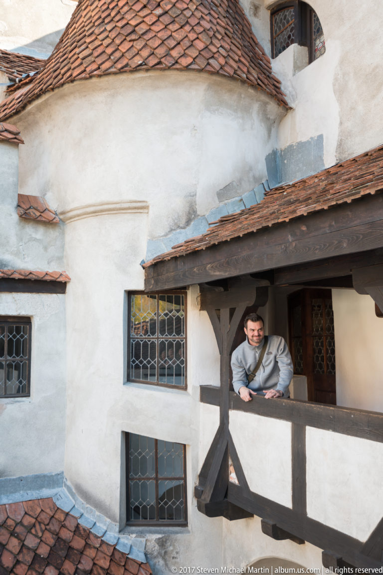 Visitor leaning on a timber gallery amid white masonry walls, leaded windows, and red-tiled roofs.