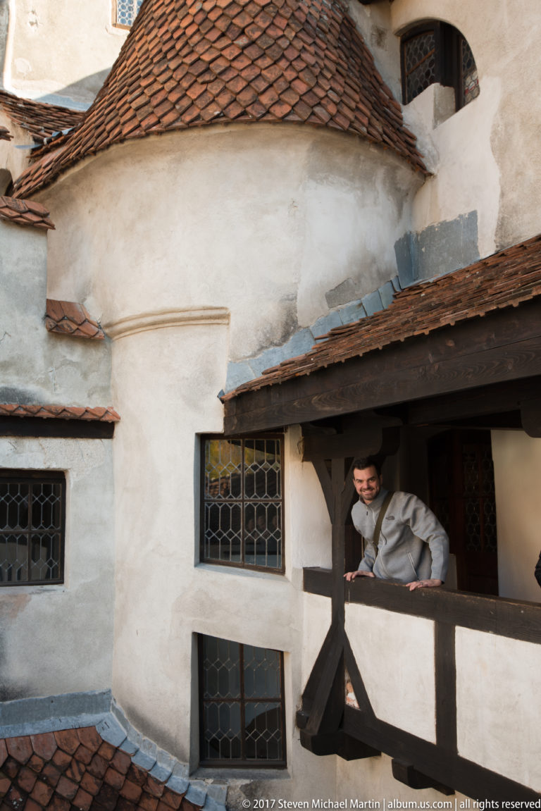 Sunlit timber gallery with a visitor, white plaster walls, small leaded windows, and tiled roofs.