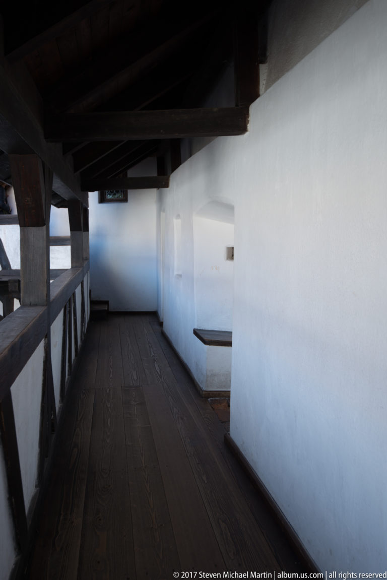 Covered wooden gallery with plank floor, timber railing, and white-plastered wall beneath dark beams.