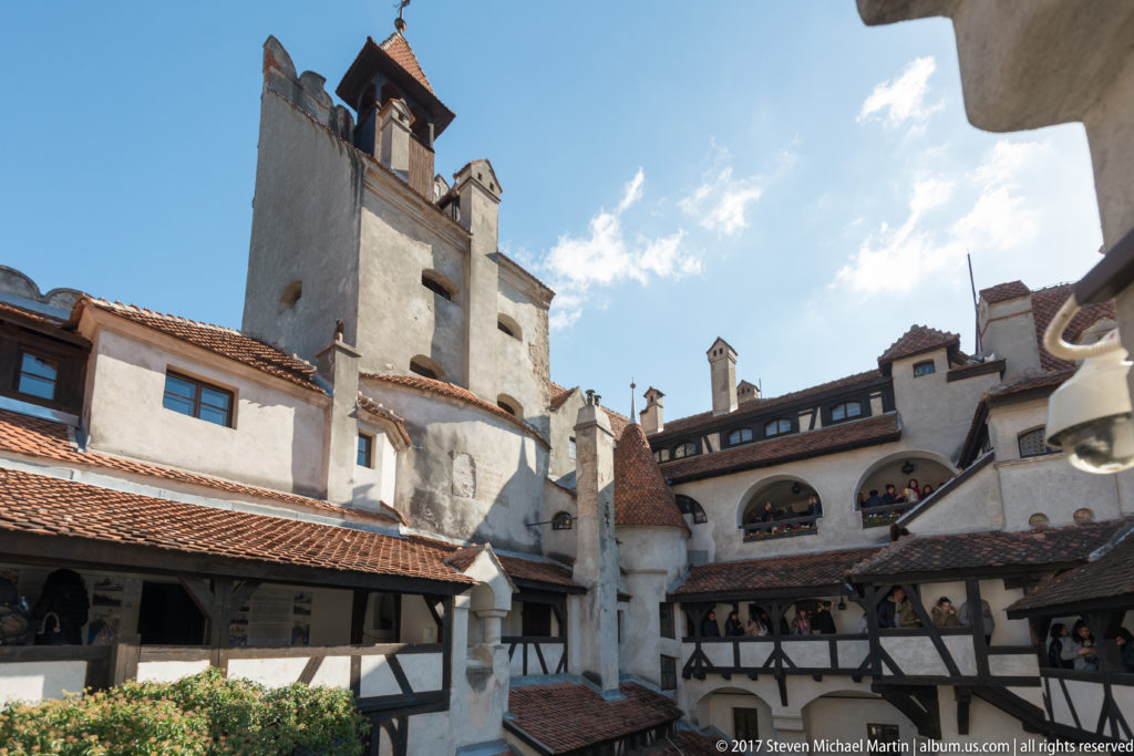 Inner courtyard with tall tower, white walls, red-tiled roofs, chimneys, arches, and timber galleries.