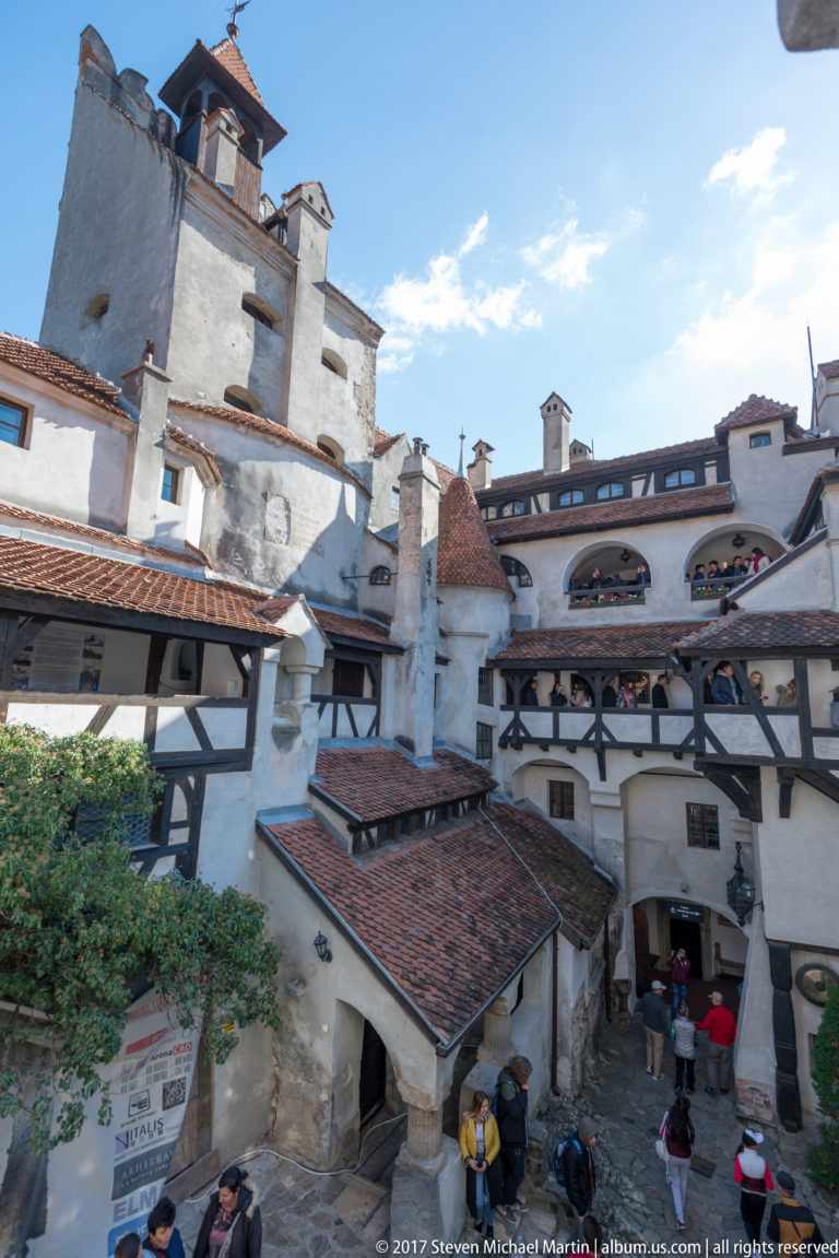 Elevated view over Bran Castle courtyard with tiled roofs, timber galleries, chimneys, and visitors below.