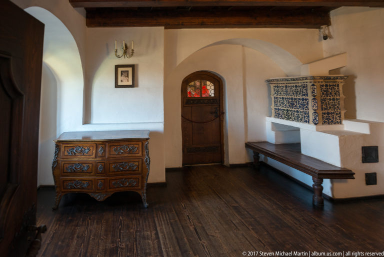 White-walled castle room with painted chest, bench, arched doorway, and blue-and-white tiled stove.