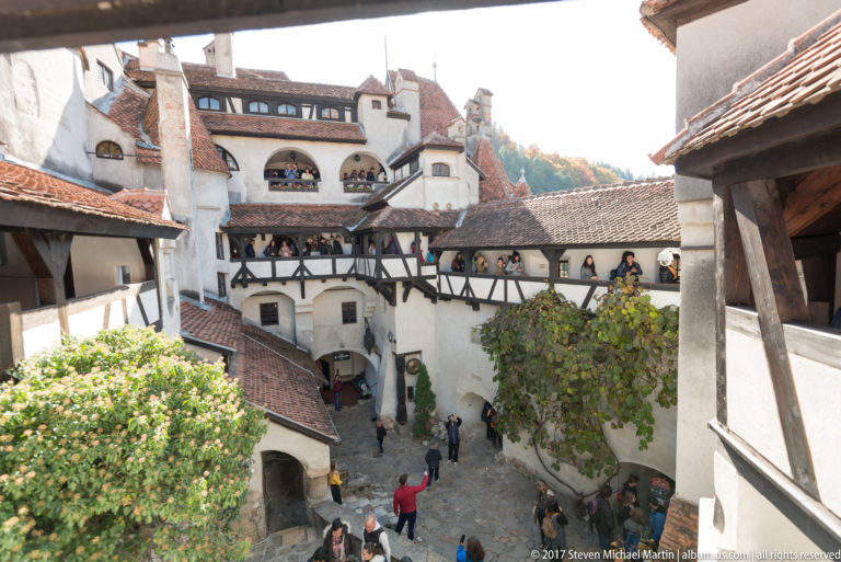 High view into stone-paved courtyard with visitors, white walls, red roofs, and crossing timber galleries.
