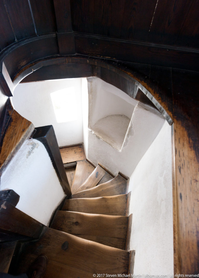 Steep wooden stairs curve downward between white plaster walls and heavy dark timber beams inside Bran Castle.
