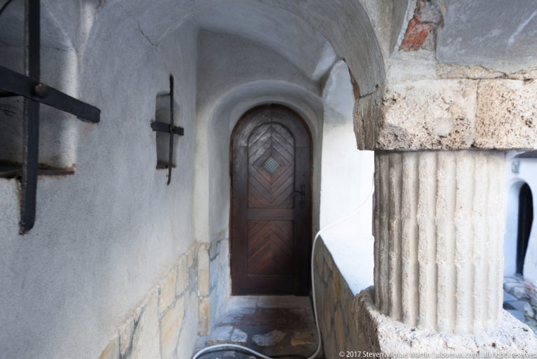 Arched paneled wooden door beside a carved stone column and exposed masonry in a narrow Bran Castle corridor.