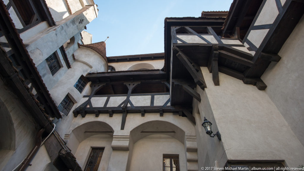 Upward view of white plaster walls, timber-framed galleries, windows, and deep eaves around Bran Castle’s courtyard.