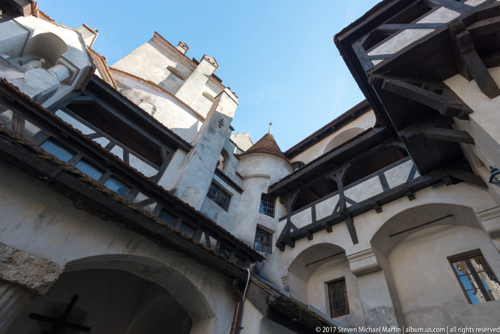 Oblique courtyard view of a pale tower, rounded turret, timber galleries, and arched openings at Bran Castle.