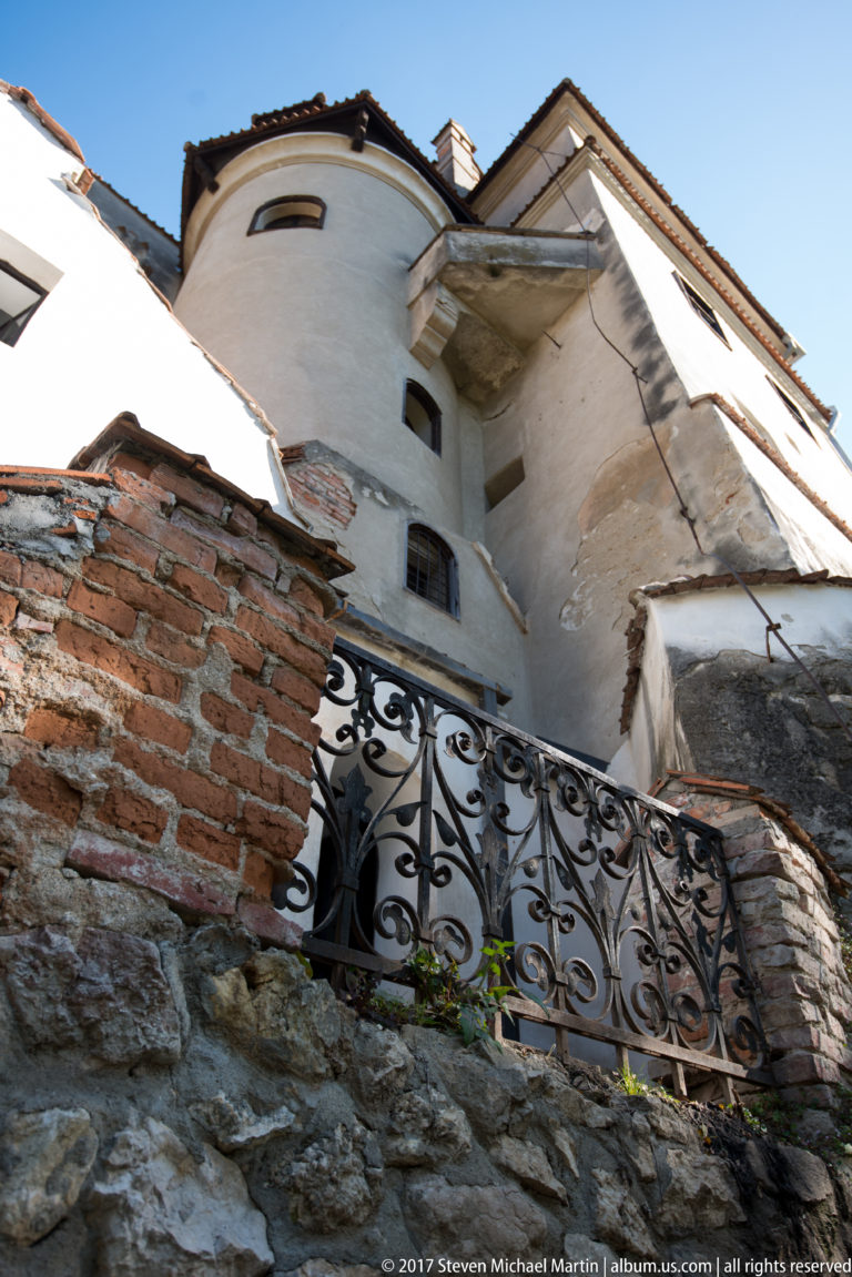 Upward exterior view of pale castle walls, rough stonework, exposed brick, small windows, and wrought-iron railing.