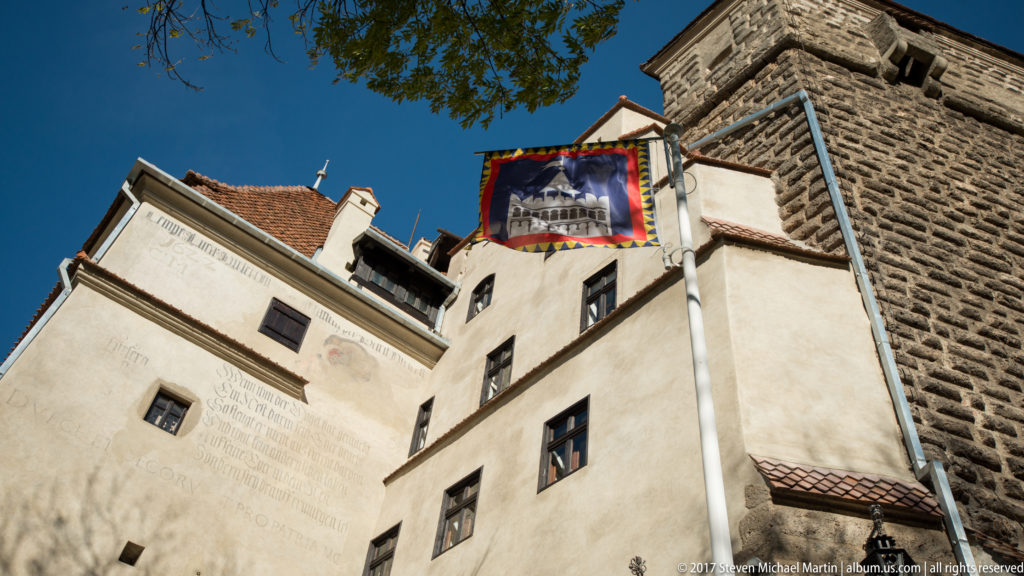 White castle walls and square stone tower with a hanging banner beneath a clear blue sky at Bran Castle.