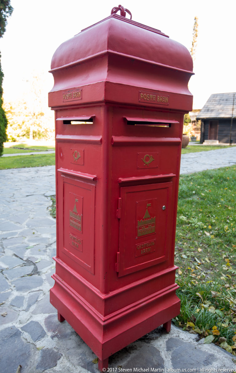 Red freestanding Romanian postal box with decorative paneling beside a paved area and grass near Bran Castle.