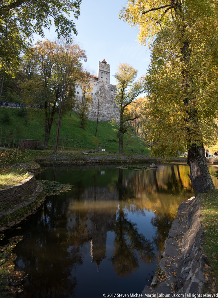 Bran Castle rises beyond a reflective pond and autumn trees, with its pale tower visible on the wooded slope.