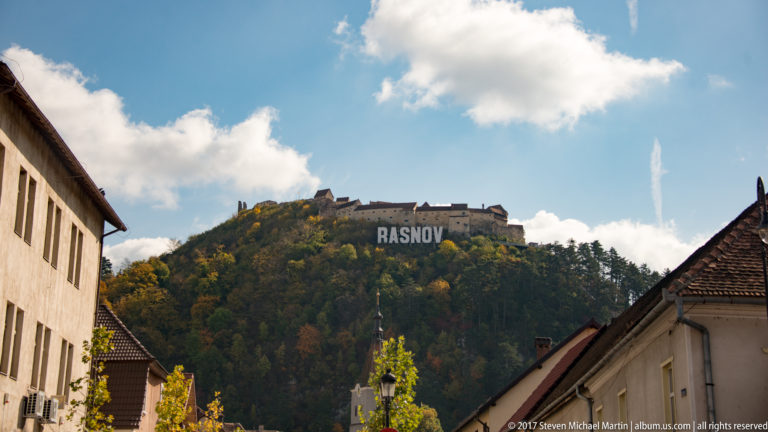 Râșnov Fortress on a wooded hill above town rooftops, with the large RASNOV sign visible on the slope.