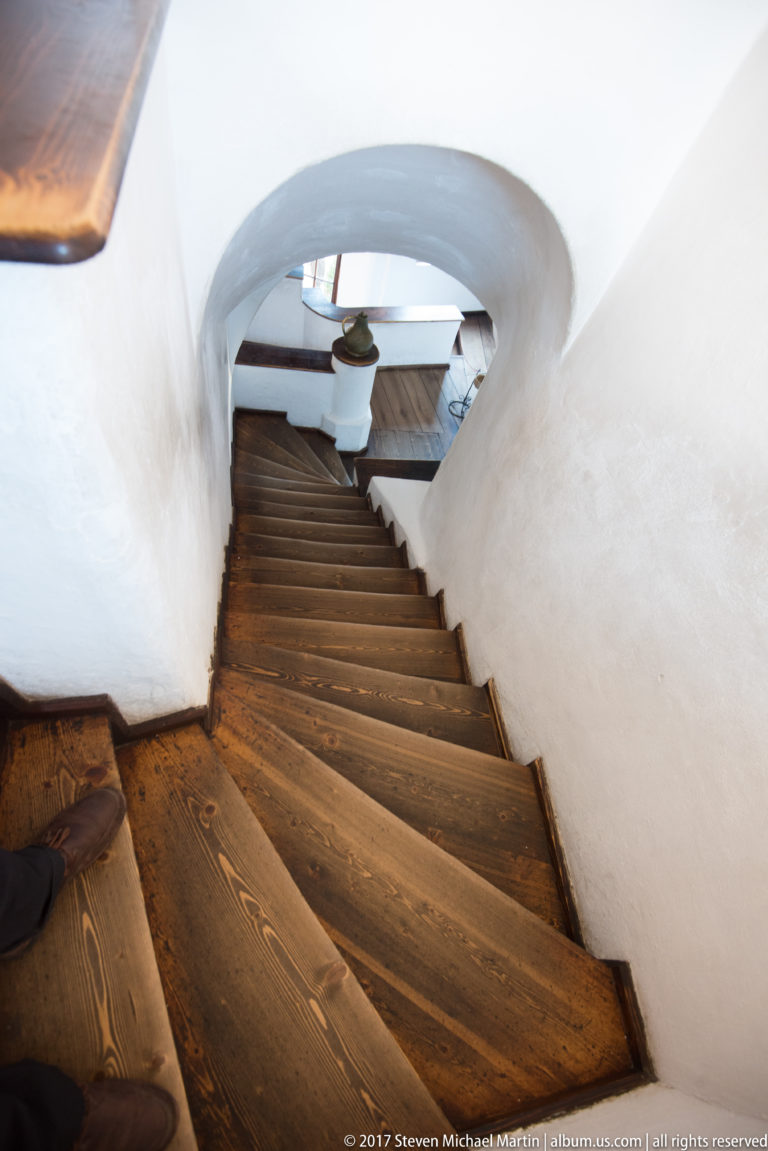 Wooden stairs descend beneath a low plastered arch toward a lower interior level.