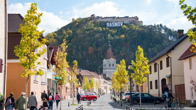 Râșnov street with pedestrians and a white church tower, while the fortress and RASNOV sign rise on the hill beyond.
